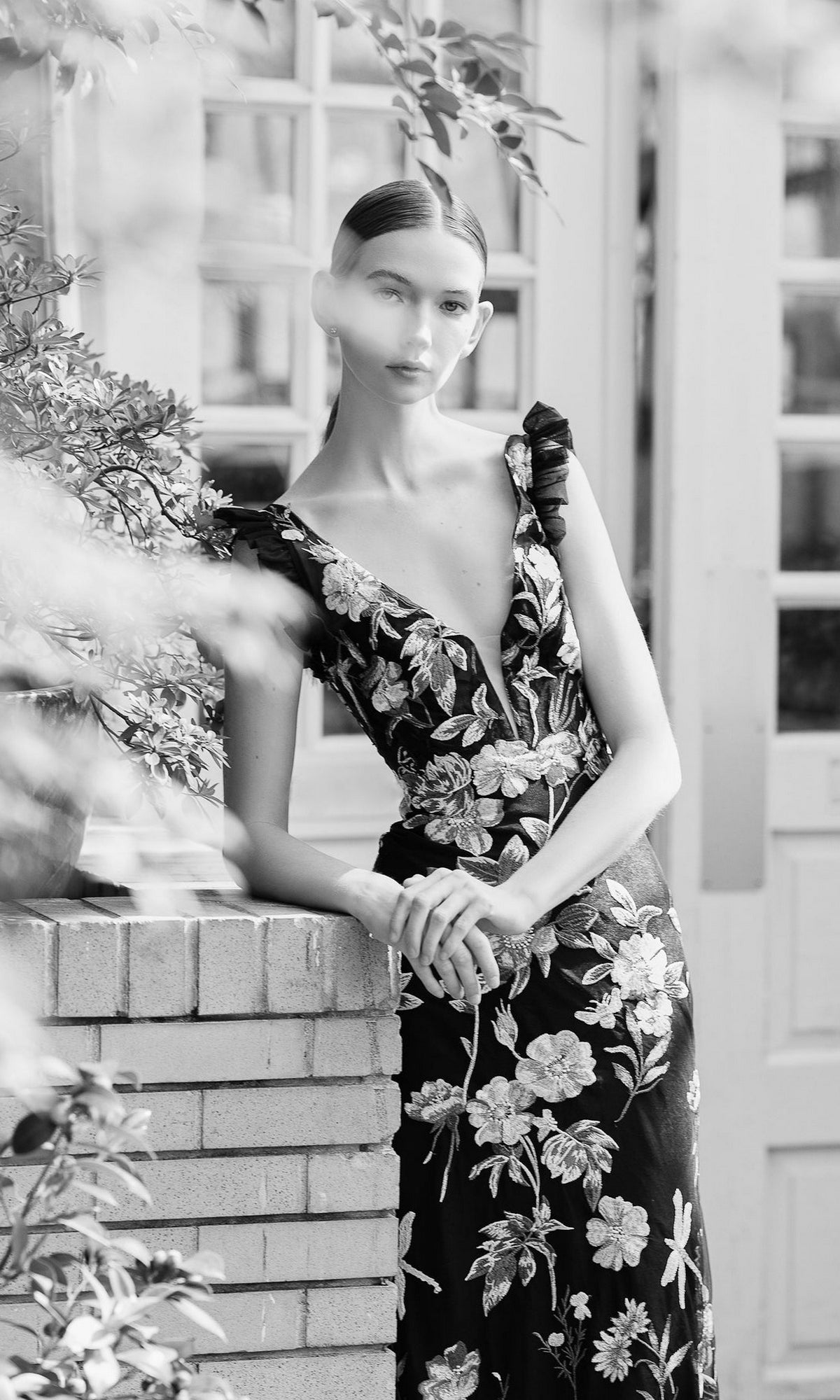 A woman wearing the Long Formal Dress 68273 by Mac Duggal, featuring black floral embroidery and a plunging v-neckline, leans against a brick wall outside with her hands clasped, gazing at the camera in this black-and-white image.